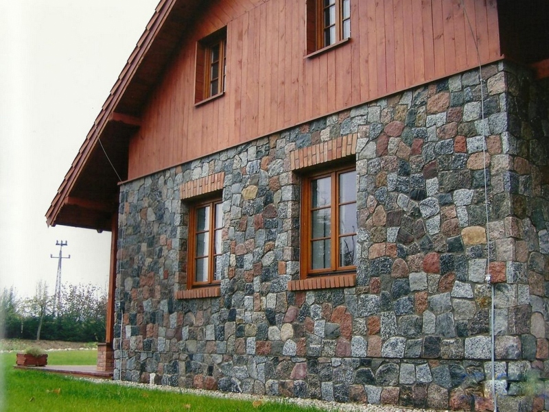 Dom z elewacją z nieregularnych, szarych i czerwonych kamieni, drewniana góra i obramowania okien, widok z boku na tle zielonego trawnika.