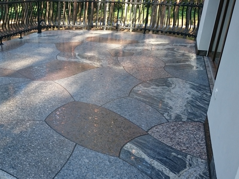 Wzorzysta, polerowana posadzka tarasu z nieregularnych płyt kamiennych w odcieniach szarości, brązu i błękitu, otoczona metalową balustradą i drzewami w tle.