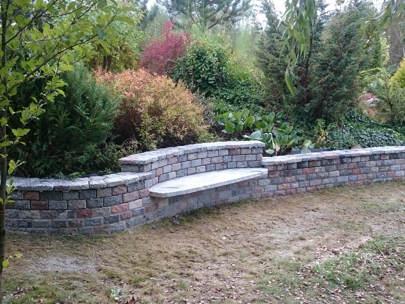 Ogród z niskim murem oporowym z kostki w odcieniach szarości i różu, zakończonym siedziskiem z jasnego kamienia, na tle zieleni i krzewów.