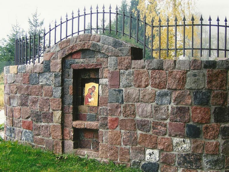 Półokrągły murek z nieregularnych, różnokolorowych kamieni granitowych z wnęką, w której umieszczono ikonę religijną; nad murkiem widoczne metalowe ogrodzenie z ostrymi zakończeniami.