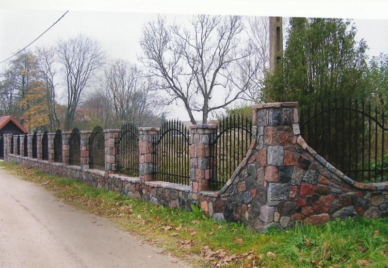 Kamienny mur z kutym żelaznym ogrodzeniem, wykończony w nieregularnym kształcie, otoczony zielenią i drzewami w jesiennej scenerii.