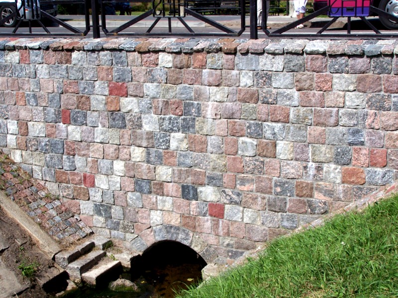 Kamienny mur z kostki granitowej w odcieniach szarości i różu, tworzący mostek nad ciekiem wodnym z widocznym tunelem przepustowym i schodkami.