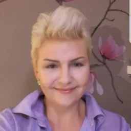 Uśmiechnięta kobieta z blond włosami w liliowej koszuli na tle ściany z motywem magnolii.