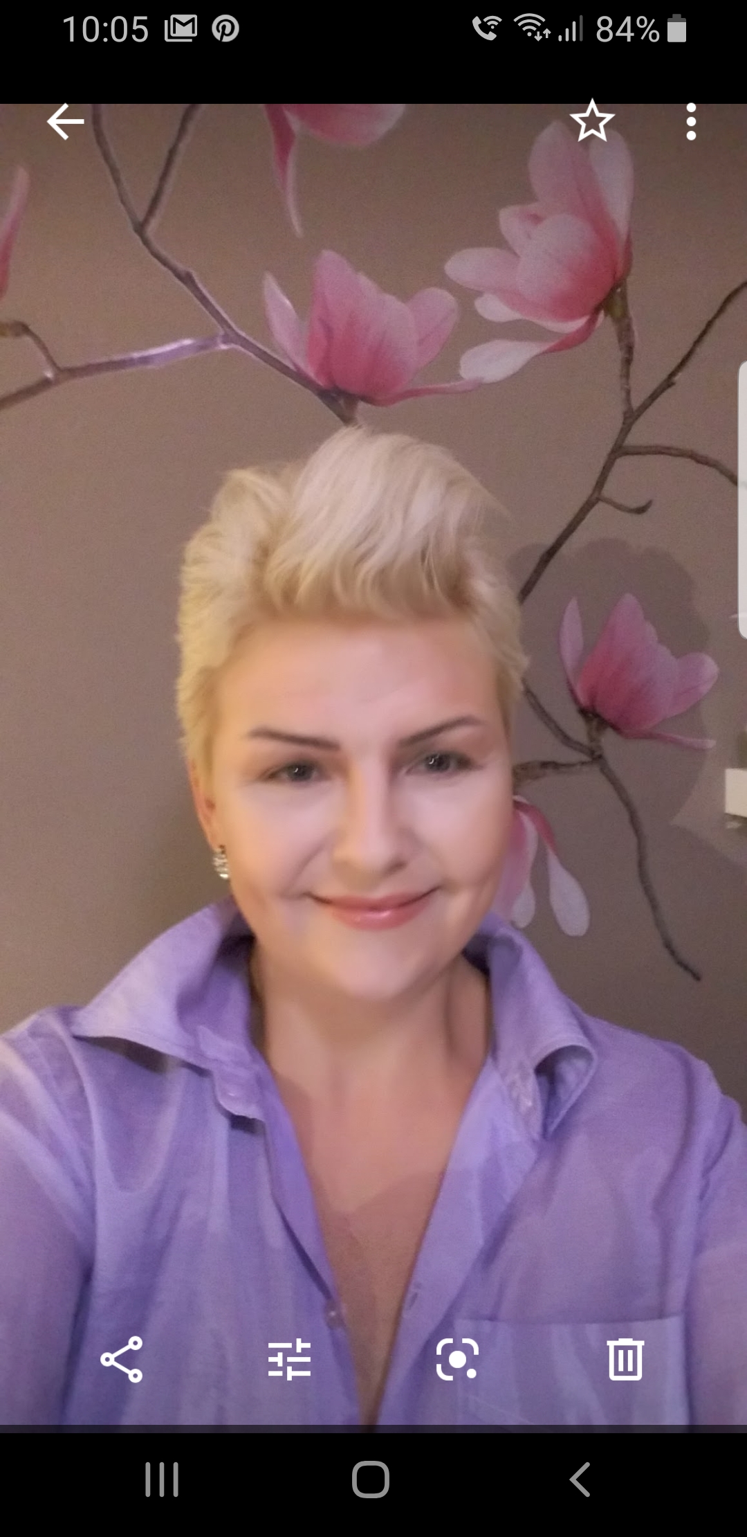 Uśmiechnięta kobieta z blond włosami w liliowej koszuli na tle ściany z motywem magnolii.