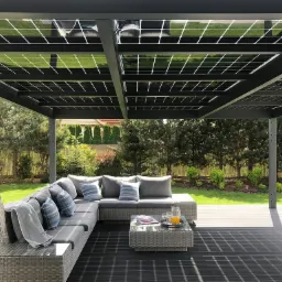 Pergola z panelami słonecznymi nad strefą relaksu z narożnikiem, stolikiem kawowym i wiszącym fotelem na tle zielonego ogrodu.