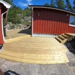 Nowa drewniana platforma tarasowa z zaokrąglonymi krawędziami, przylegająca do czerwonych budynków na skalistym terenie.