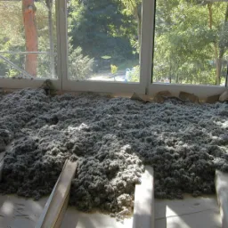 Niedokończona izolacja poddasza wełną celulozową w Sopotni Małej, widoczne drewniane legary i okno z widokiem na zieleń.