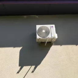 Zewnętrzna jednostka klimatyzacji marki Gree zamontowana na jasnej, teksturowanej ścianie budynku, oświetlona mocnym słońcem rzucającym wyraźny cień urządzenia na elewację.