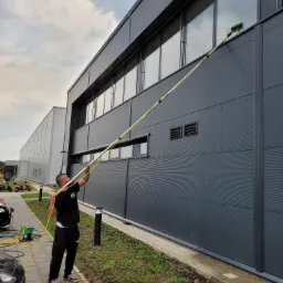 Pracownik myje okna na wysokości w nowoczesnym biurowcu z szarej blachy falistej, używając teleskopowej lancy z zieloną szczotką, widoczny wąż i zaparkowane skutery.
