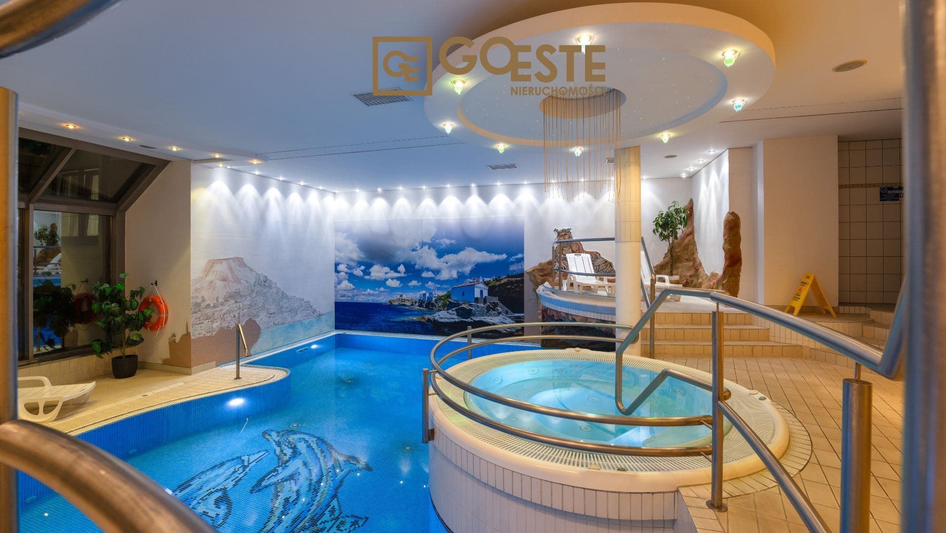 Kryty basen z jacuzzi, mozaiką delfina na dnie, fototapetą z motywem morskim i dekoracyjnym oświetleniem sufitowym w pomieszczeniu z logo agencji nieruchomości.