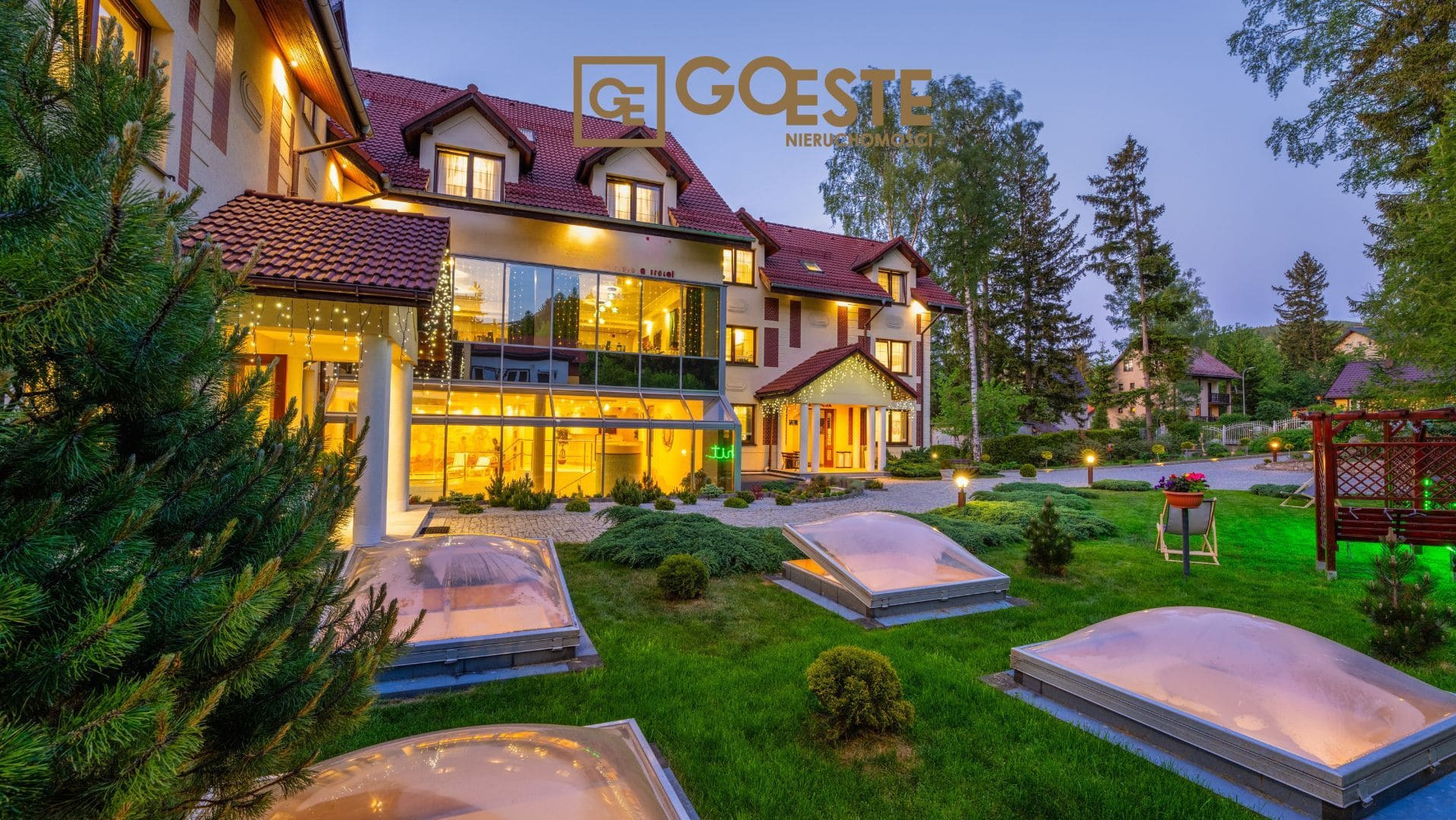 Elegancki, oświetlony budynek z przeszkloną fasadą i logo firmy nieruchomości na tle wieczornego nieba, z trawnikiem i świetlikami w pierwszym planie.