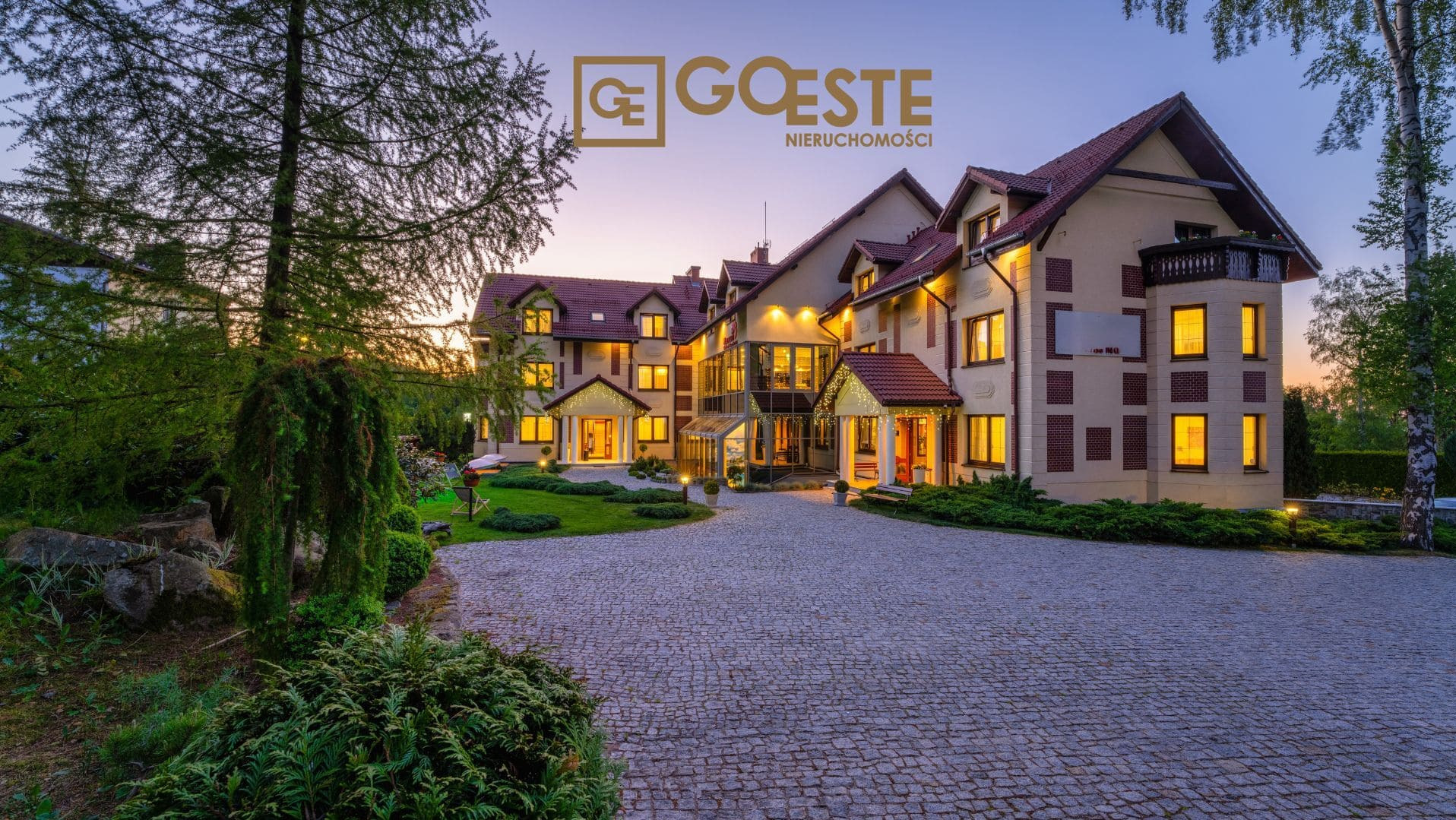 Elegancki budynek z podświetlonymi oknami i reprezentacyjnym podjazdem z kostki brukowej, logo firmy nieruchomości na fasadzie, widok o zmierzchu.