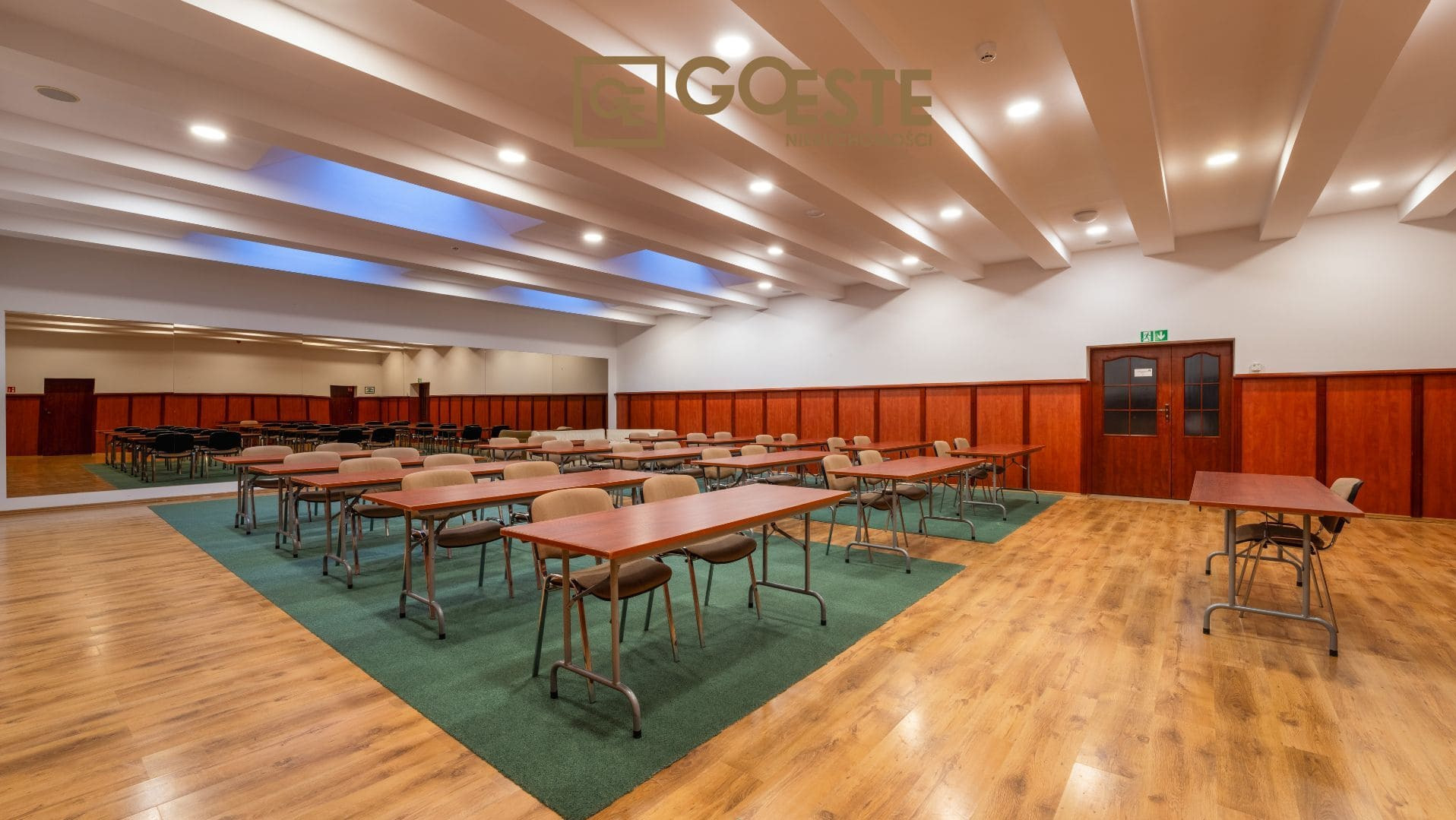 Przestronna sala konferencyjna z drewnianą podłogą, stołami i krzesłami ustawionymi w rzędach, zielonym dywanem i dużym lustrem na ścianie, oświetlona jasnymi lampami sufitowymi.