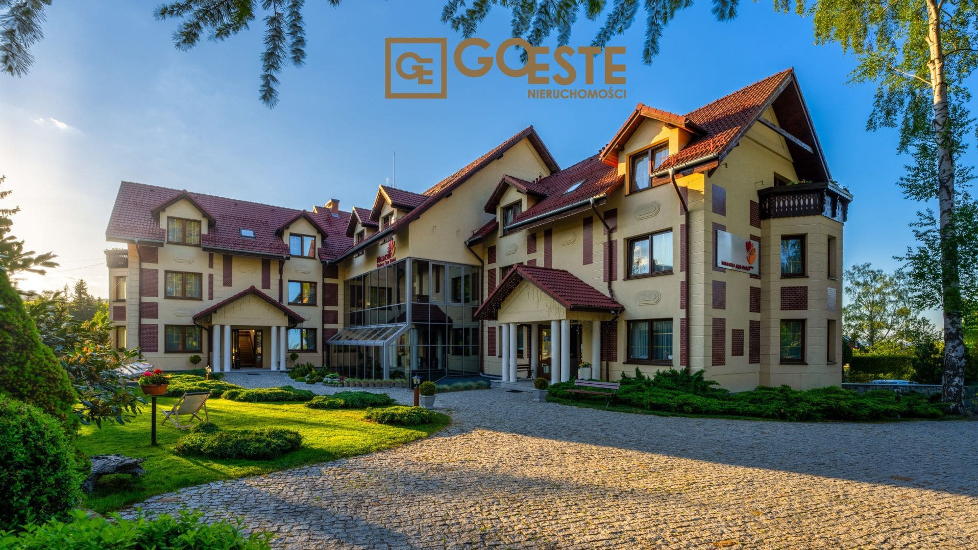 Elegancki budynek agencji nieruchomości z beżową elewacją, czerwoną dachówką i przeszklonym wejściem, otoczony zadbanym trawnikiem i brukowanym podjazdem w słoneczny dzień.