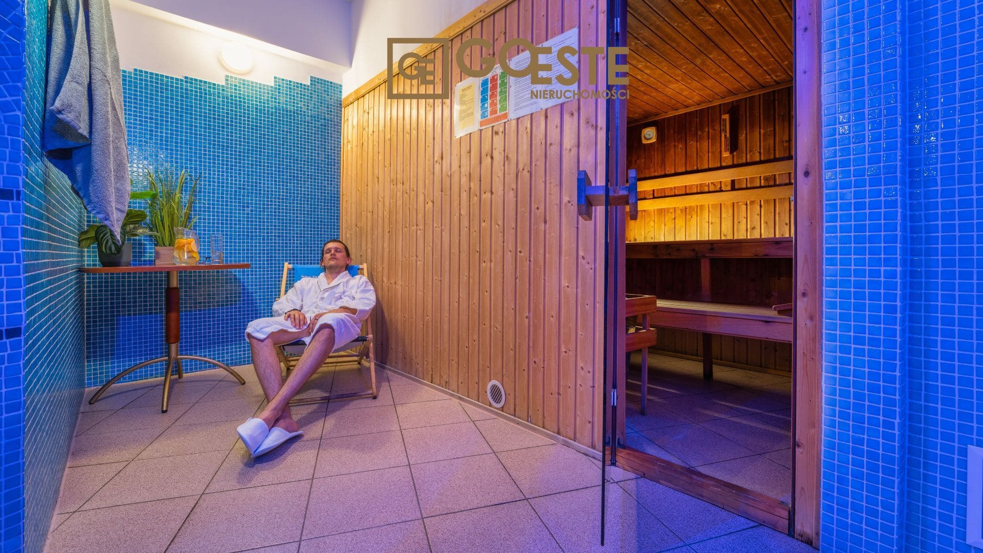 Relaksujący mężczyzna w szlafroku siedzi na krześle w pomieszczeniu z sauną, niebieską mozaiką i drewnianą ścianą z logo agencji nieruchomości.