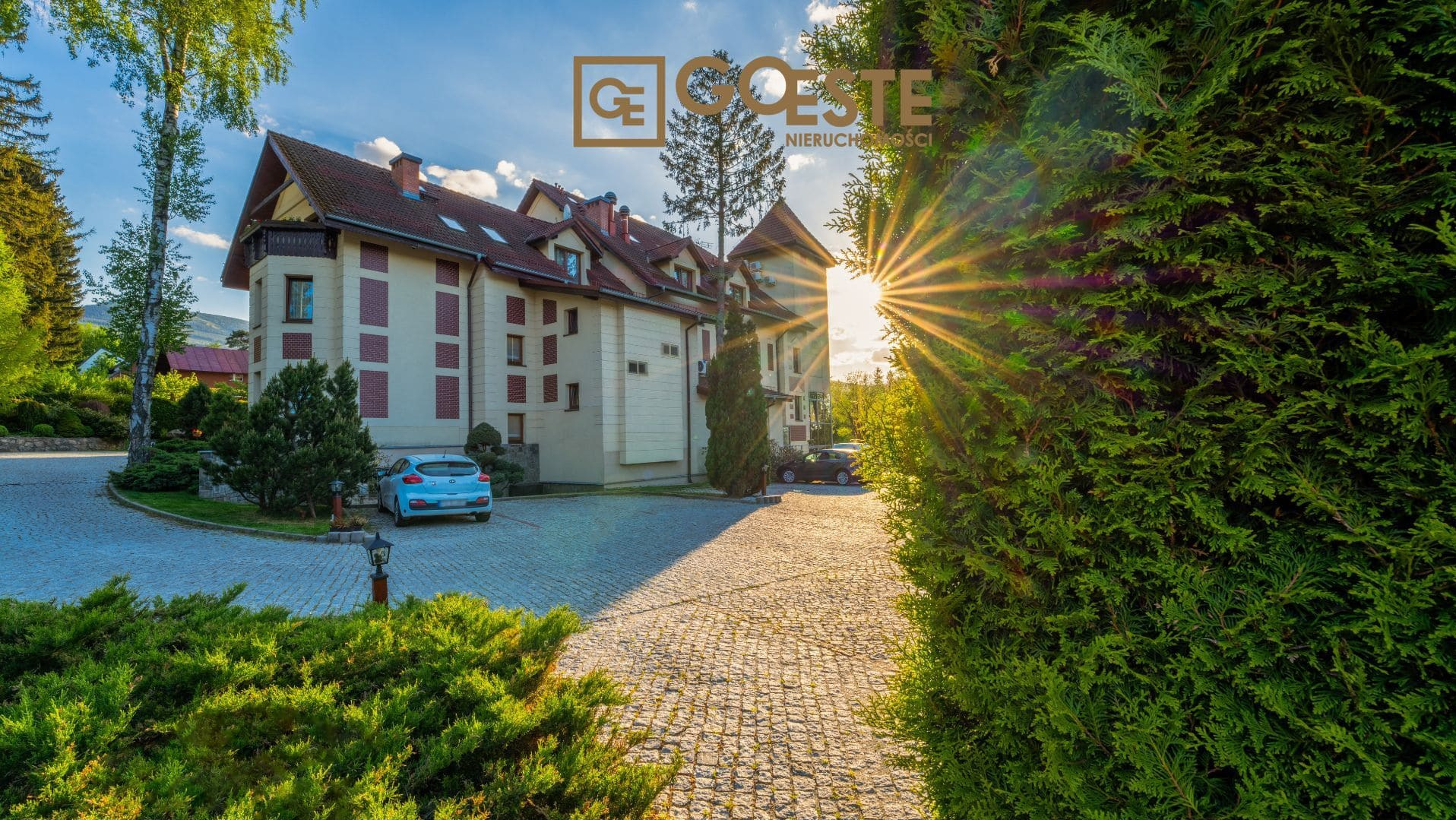 Elegancki budynek z dwuspadowym dachem i wieżyczką, kremowa elewacja z ceglanymi detalami, samochody na brukowanym podjeździe, logo agencji nieruchomości w górnej części kadru, promienie słońca...