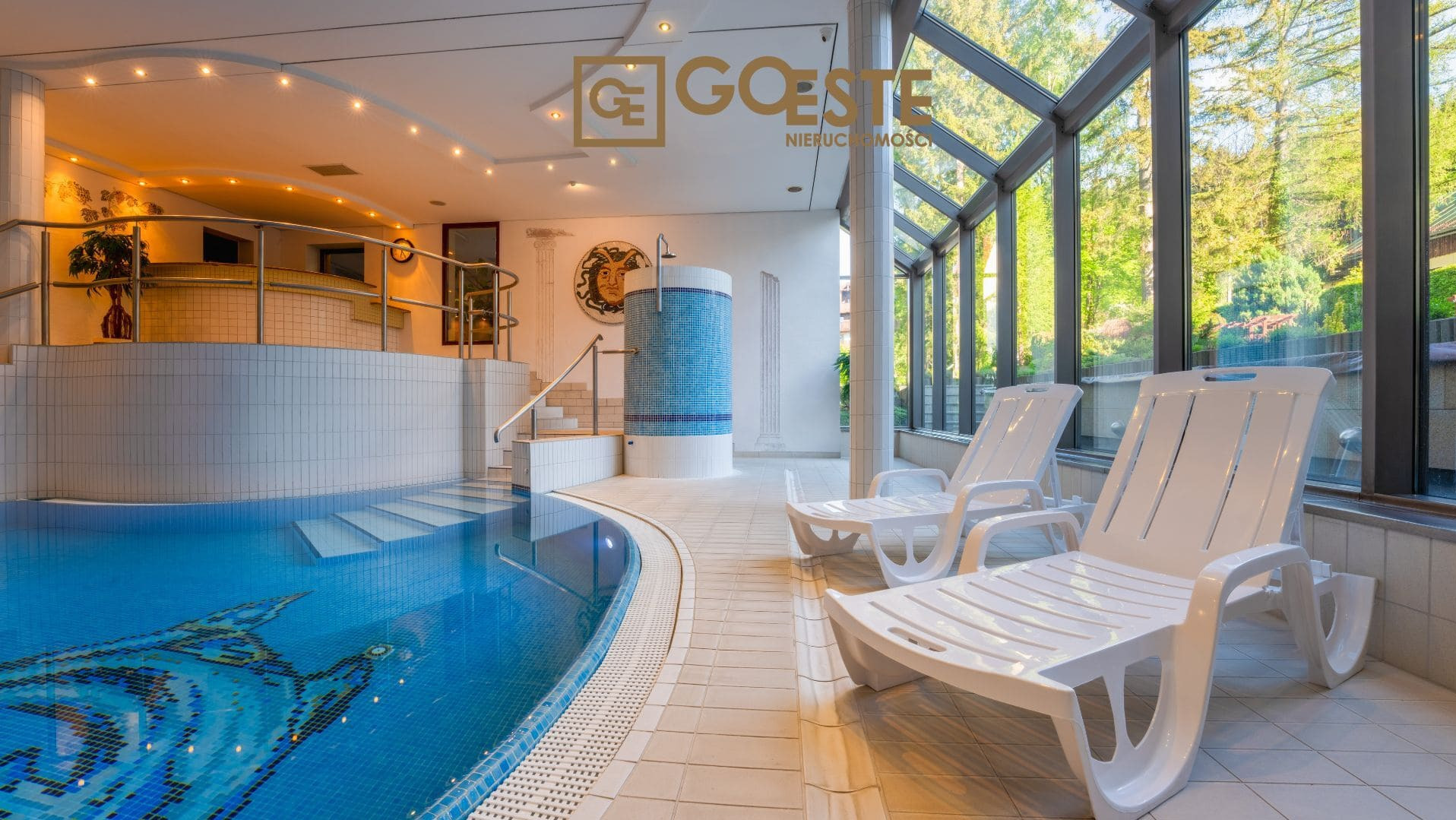 Kryty basen z mozaiką przedstawiającą rybę, białe leżaki przy przeszklonej ścianie z widokiem na zieleń, oraz logo 'GOESTE Nieruchomości' w górnej części zdjęcia.