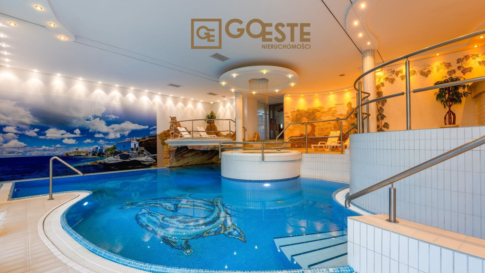 Luksusowy basen kryty z mozaiką delfina na dnie, dekoracyjnym oświetleniem i muralem z motywem śródziemnomorskim na ścianie. Widoczne logo firmy Goeste Nieruchomości.