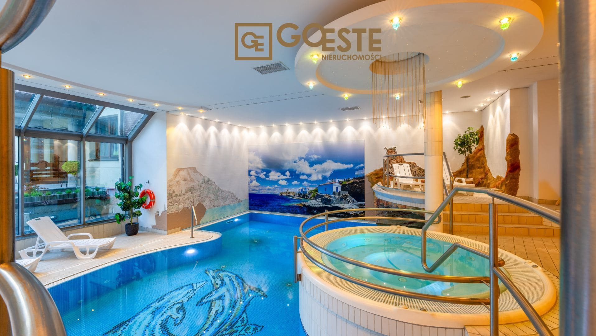 Luksusowy kryty basen z jacuzzi, mozaiką z motywem delfina i dekoracyjną ścianą z motywami krajobrazowymi, oświetleniem punktowym i roślinami doniczkowymi.