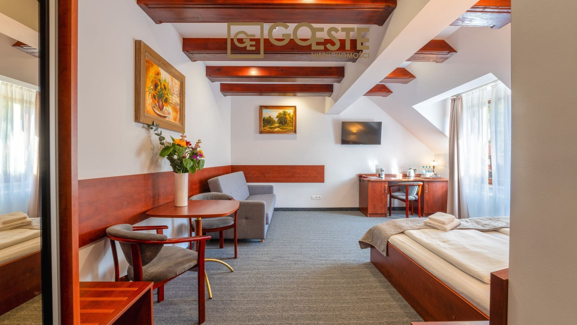 Przytulne wnętrze pokoju na poddaszu z drewnianymi belkami stropowymi, obrazami na ścianach, sofą, stołem z kwiatami, biurkiem i łóżkiem z pościelą.