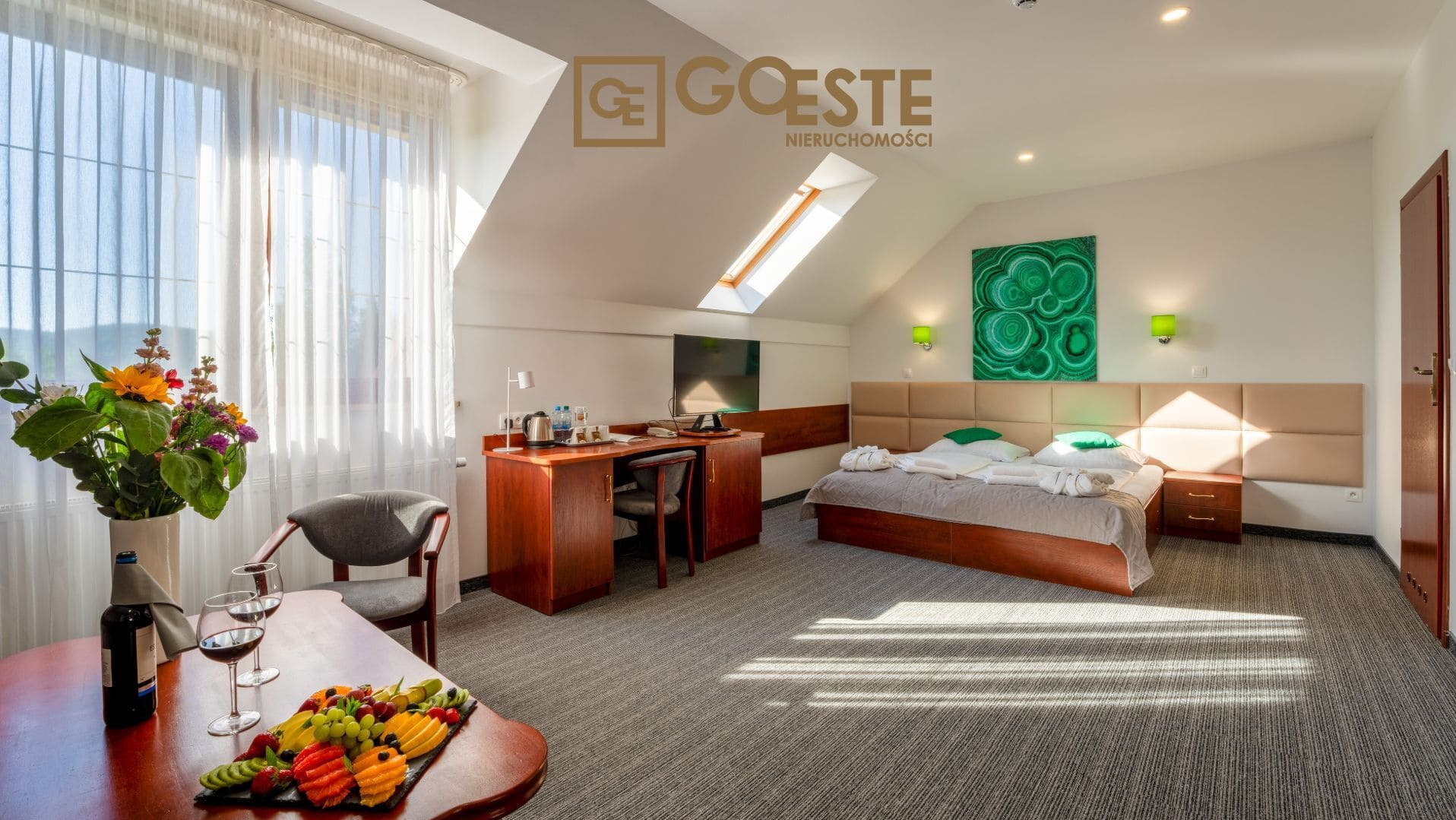 Jasny pokój hotelowy na poddaszu z biurkiem, łóżkiem, owocami, winem i kwiatami. Logo 'Goeste Nieruchomości' widoczne w górnej części zdjęcia.