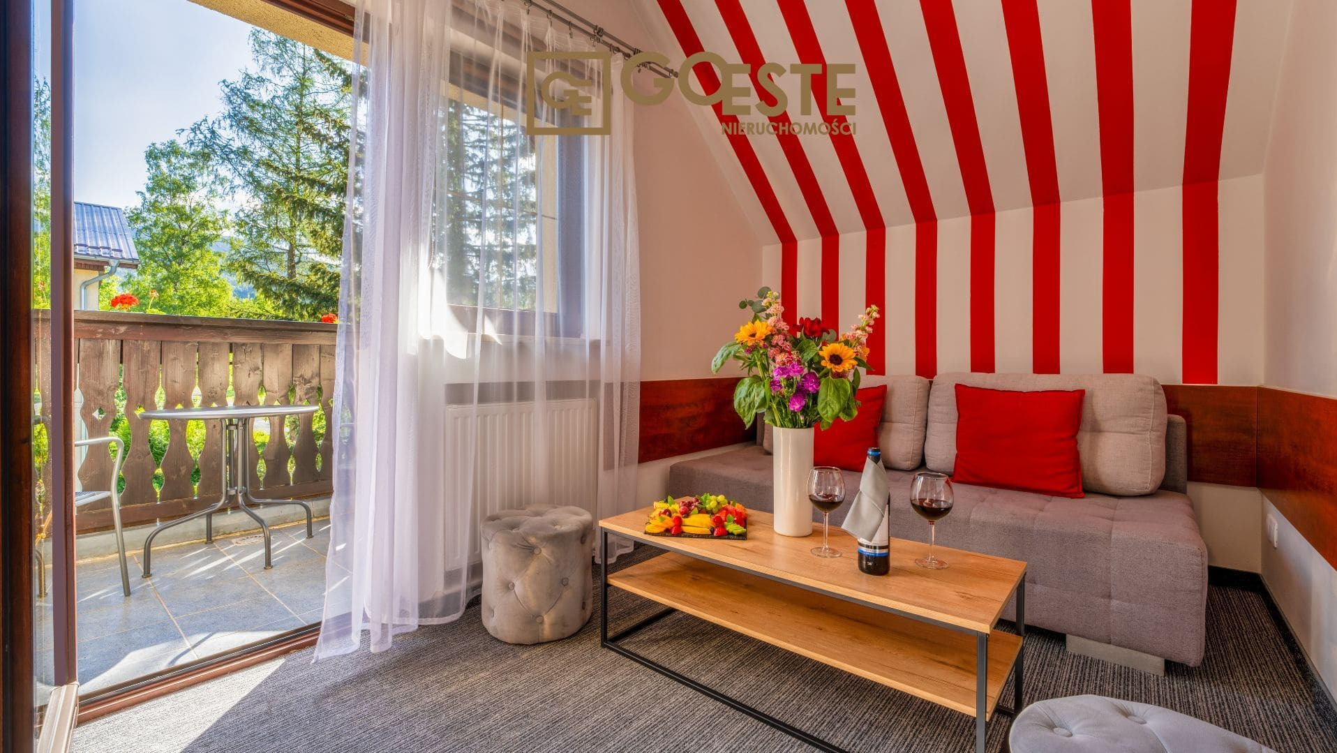 Jasne wnętrze pokoju z balkonem, stolikiem i krzesłami, kwiatami w wazonie, deską z owocami i dwoma kieliszkami wina na stoliku kawowym, ściany w biało-czerwone pasy.