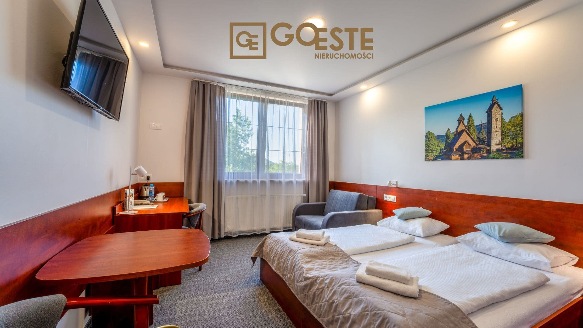 Jasny pokój hotelowy z dwoma łóżkami, sofą, biurkiem i telewizorem, nad łóżkiem wisi obraz z kościołem, widoczne logo firmy Goeste Nieruchomości.