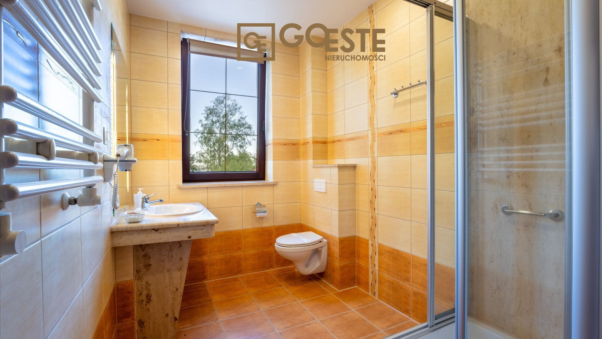 Jasna łazienka z beżowo-pomarańczowymi płytkami, umywalką na postumencie, grzejnikiem drabinkowym i kabiną prysznicową; widok na drzewo za oknem, logo agencji nieruchomości w górnej części zdjęcia.