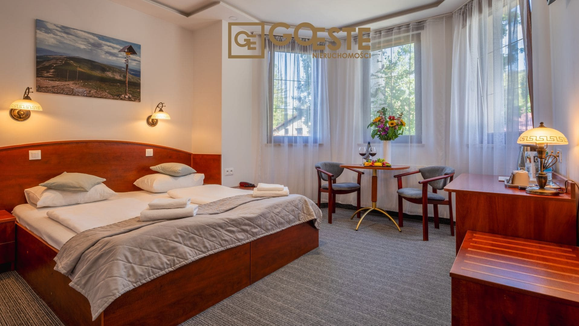 Pokój hotelowy z dużym łóżkiem, stolikiem z winem i owocami, oraz widokiem z okna na zieleń.