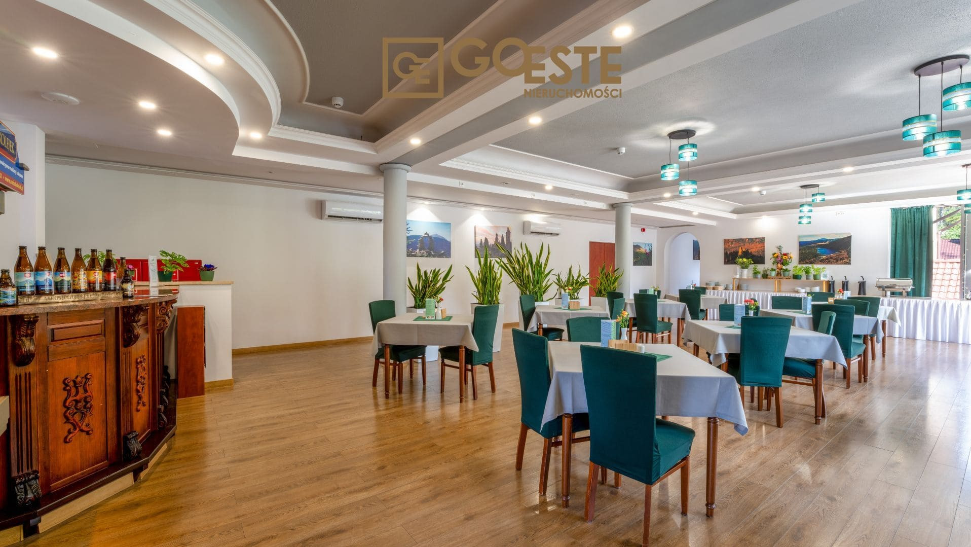 Jasna sala restauracyjna z drewnianą podłogą, nakrytymi stołami z zielonymi krzesłami, roślinami doniczkowymi i obrazami na ścianach, widoczny bar z butelkami i logo agencji nieruchomości w górnej...