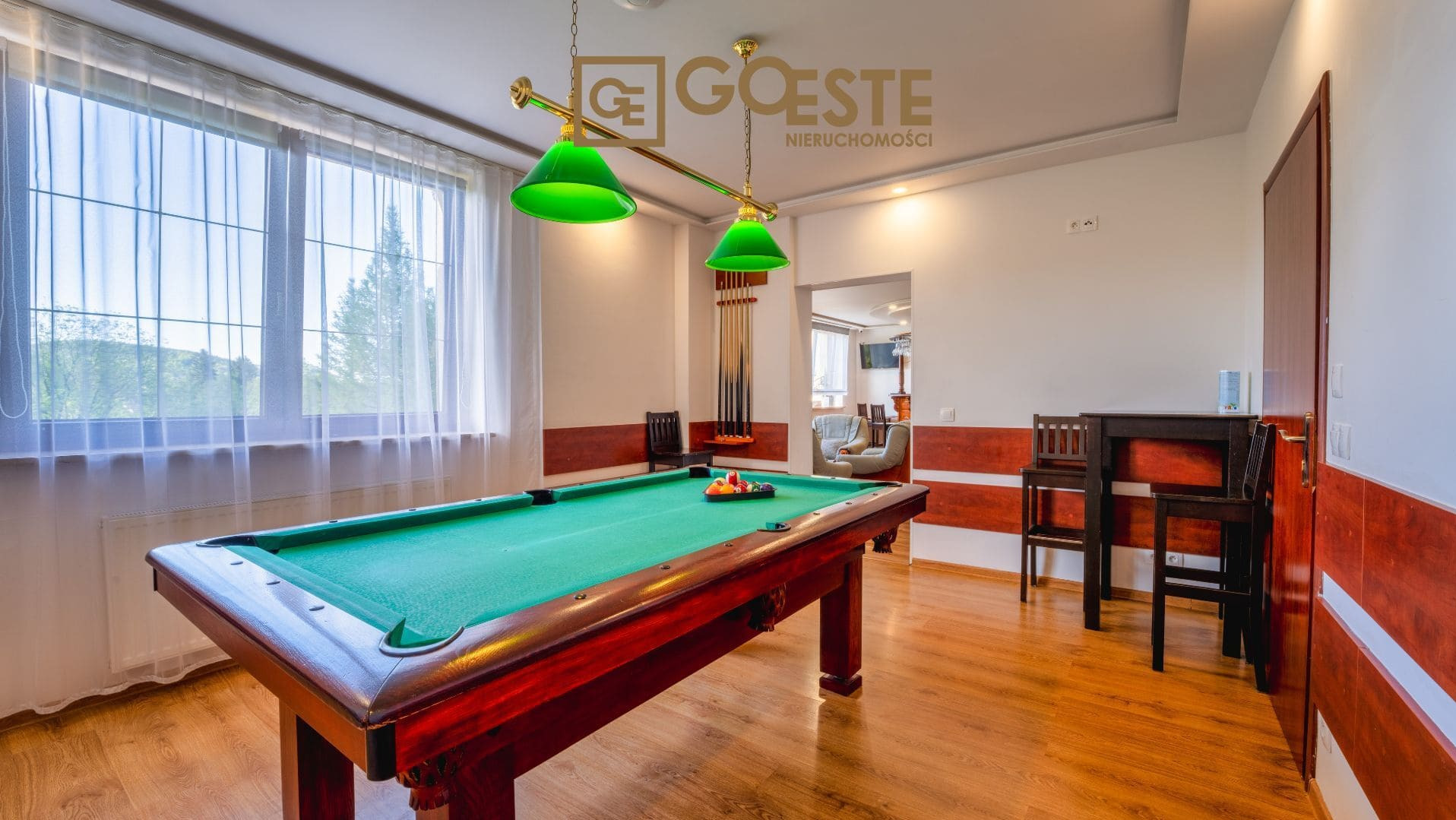 Pokój z bilardem, stół bilardowy z zielonym suknem, nad nim lampa z dwoma zielonymi kloszami, w tle logo firmy 'Goeste Nieruchomości' nad stołem.