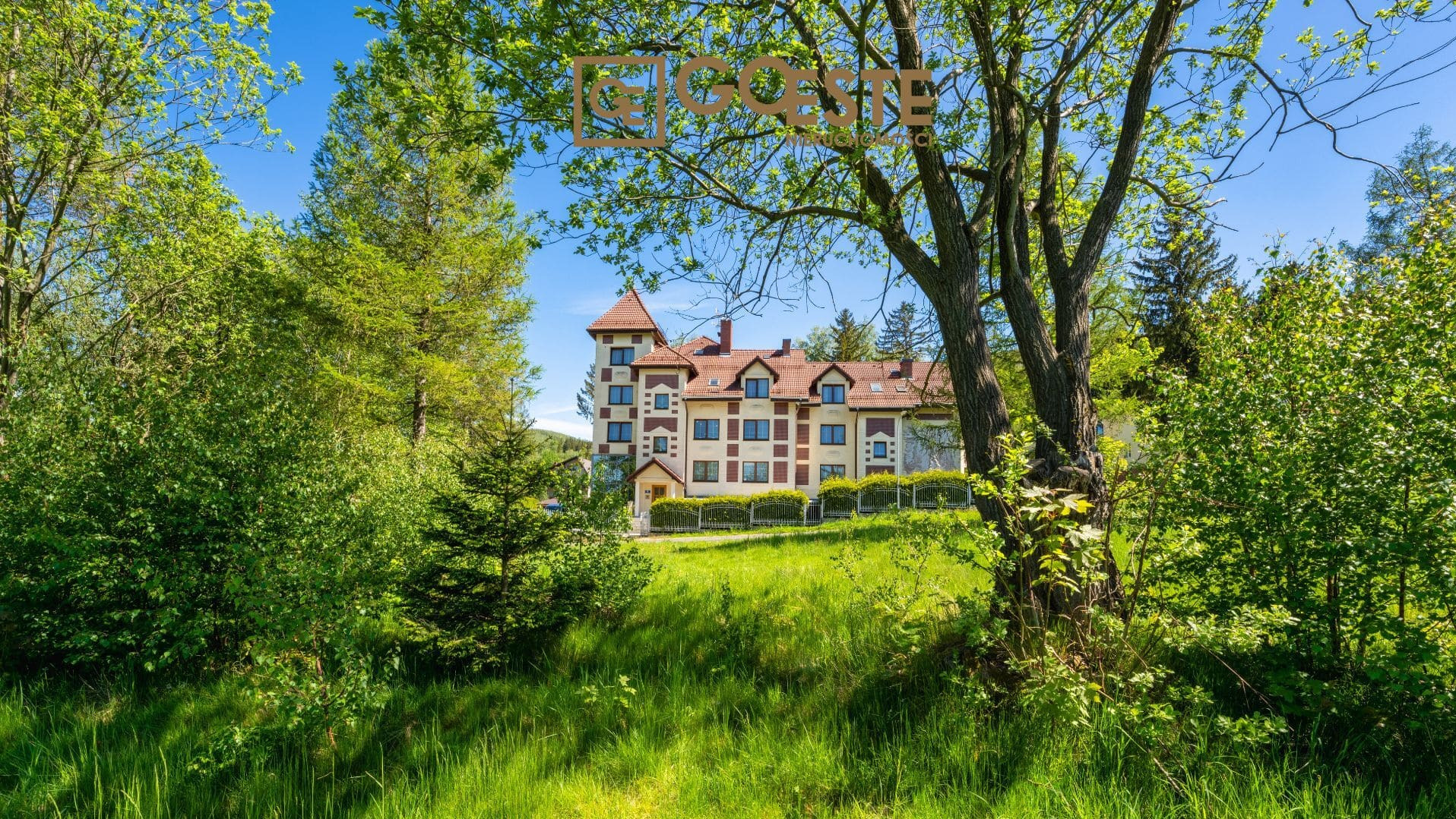 Trzypiętrowy budynek mieszkalny z czerwoną dachówką, widoczny zza drzew i bujnej zieleni w słoneczny dzień.