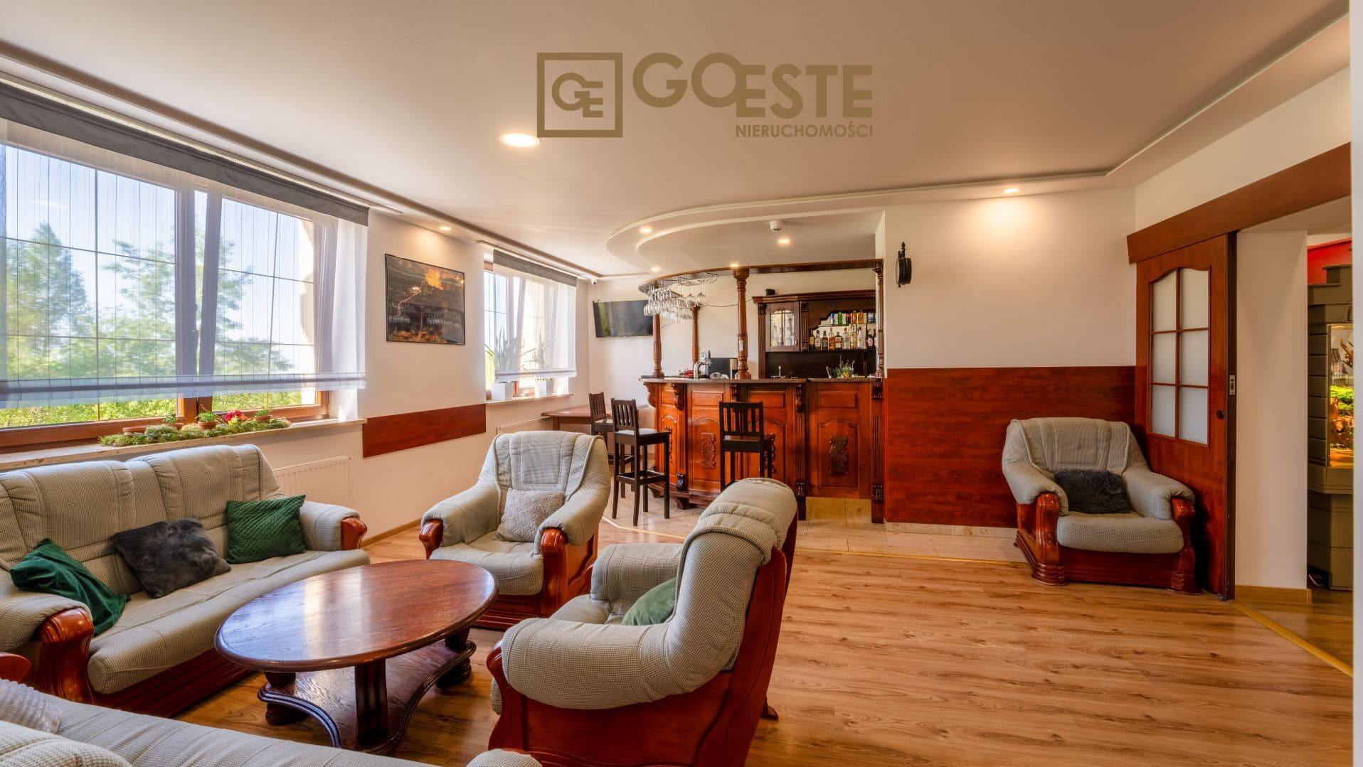 Przestronny salon z barem w stylu retro, widoczne sofy, fotele, drewniany stół i bar z wyposażeniem, logo firmy nieruchomości w górnym rogu.