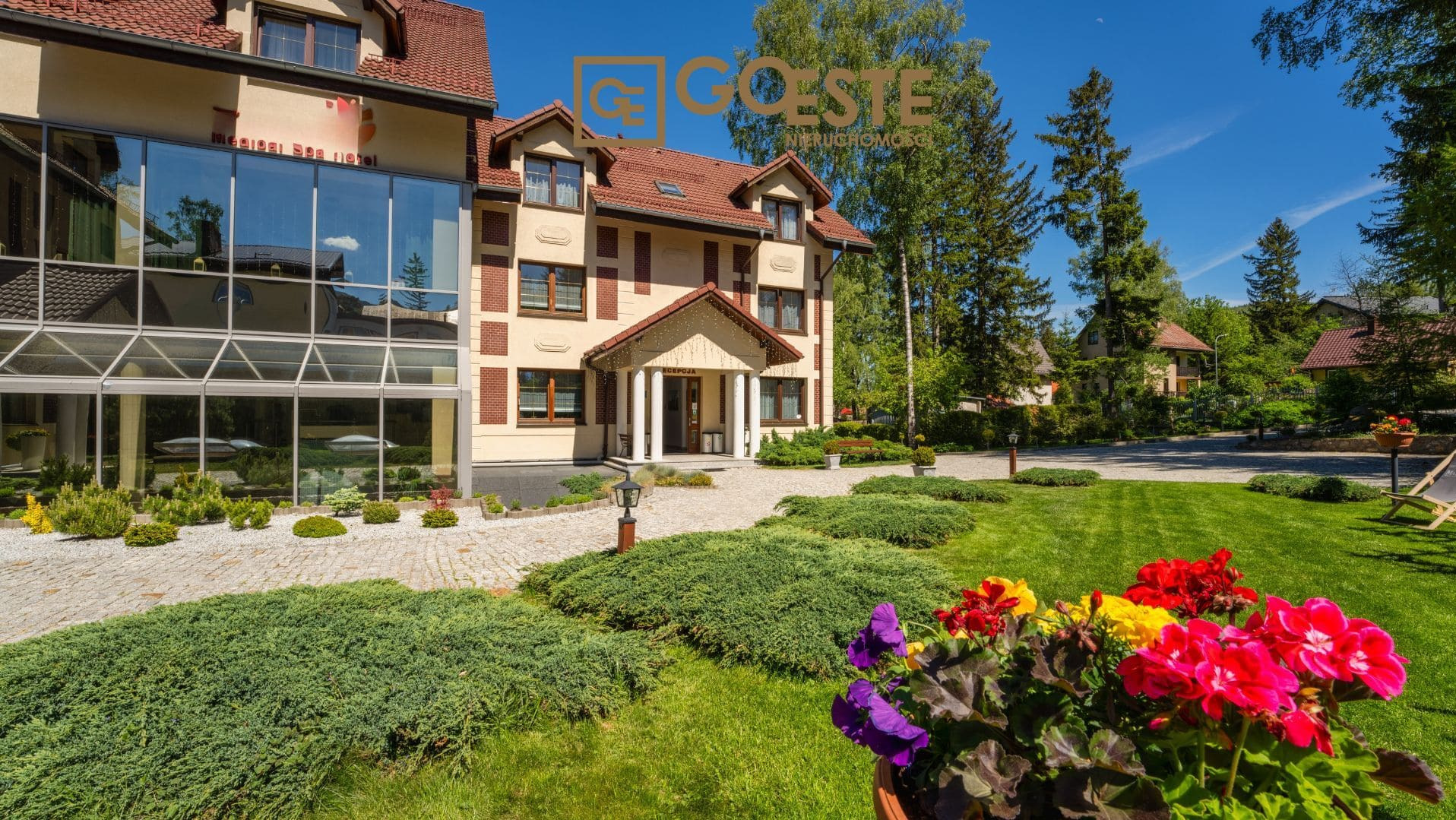 Elewacja dwukondygnacyjnego budynku z jasną fasadą i czerwoną dachówką, widoczna przeszklona część budynku i zadbany ogród z kolorowymi kwiatami w donicy na pierwszym planie.