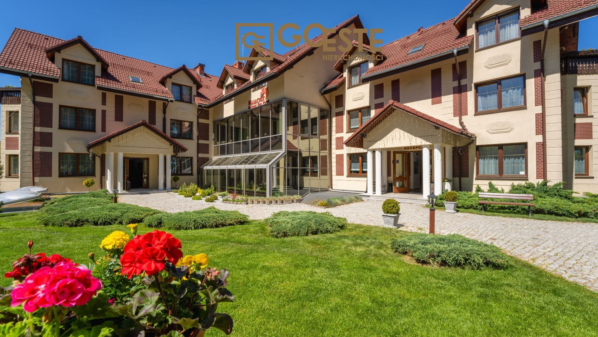 Elewacja budynku z czerwonym dachem, widok z ogrodu z rabatami kwiatowymi, przeszklone wejście do recepcji, kamienny chodnik i ławka.