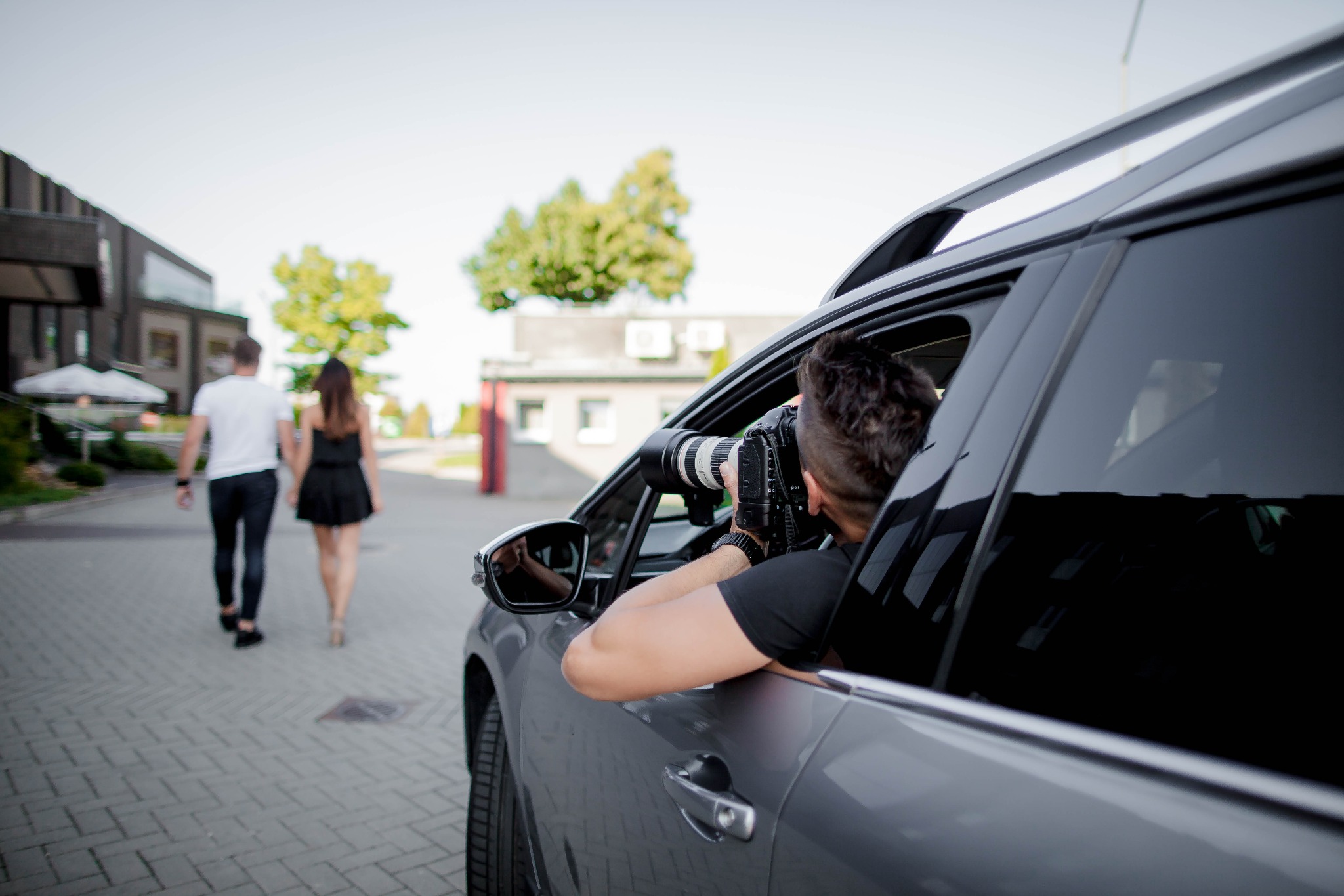 Mężczyzna w samochodzie z dużym obiektywem fotograficznym robi zdjęcia parze spacerującej po chodniku.