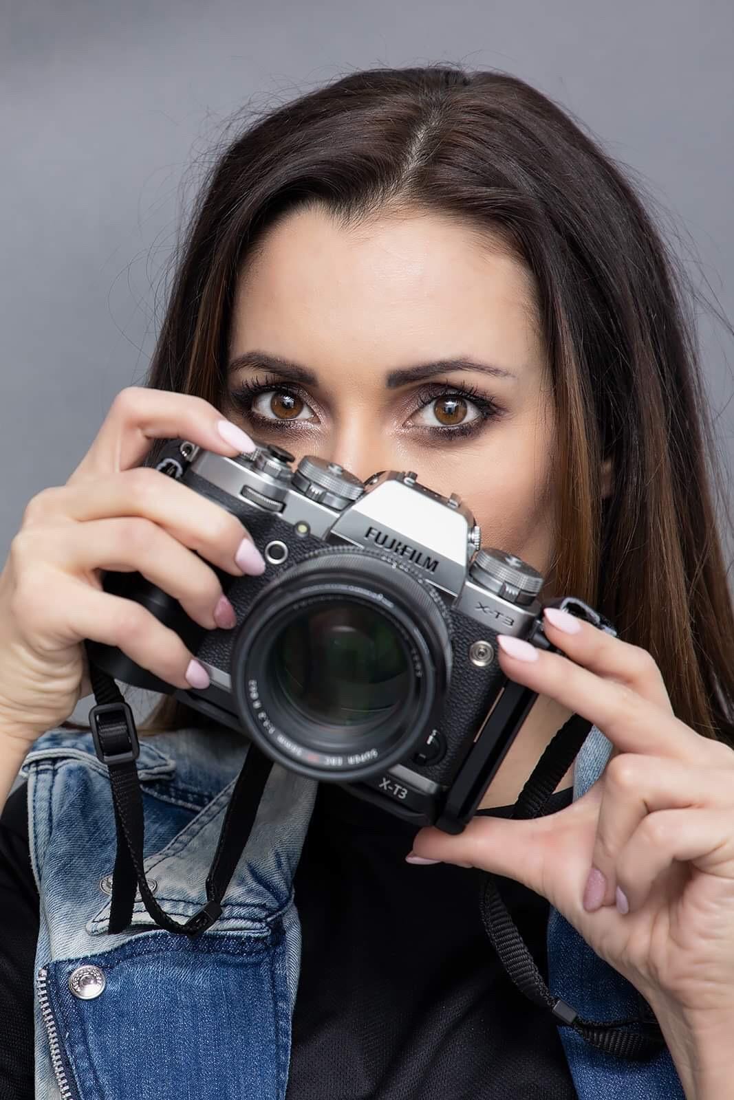 Kobieta z brązowymi oczami patrzy znad srebrno-czarnego aparatu Fujifilm X-T3 z obiektywem, trzymanego w dłoniach z jasnoróżowym manicure, ubrana w dżinsową kurtkę.