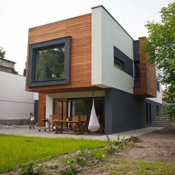 MH PRACOWNIA ARCHITEKTONICZNA MACIEJ HOFMAN - Nowoczesny budynek jednorodzinny w Poznaniu z drewnianą elewacją, dużymi oknami i tarasem z meblami ogrodowymi oraz wiszącym fotelem kokon. W tle widoczna kobieta przy grillu.