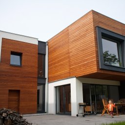 MH PRACOWNIA ARCHITEKTONICZNA MACIEJ HOFMAN - Nowoczesny budynek jednorodzinny z drewnianą elewacją i dużymi oknami, dziecko stoi na tarasie, obok stół z krzesłami i grill, widoczny stos drewna.