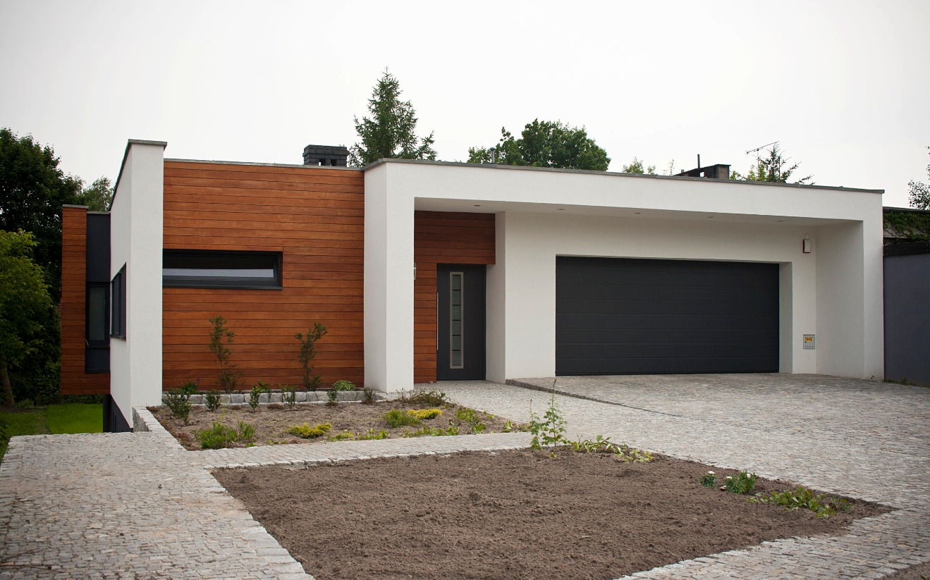 Nowoczesny budynek jednorodzinny z elementami drewnianej elewacji i garażem z bramą segmentową, otoczony kostką brukową i rabatami z młodymi roślinami.