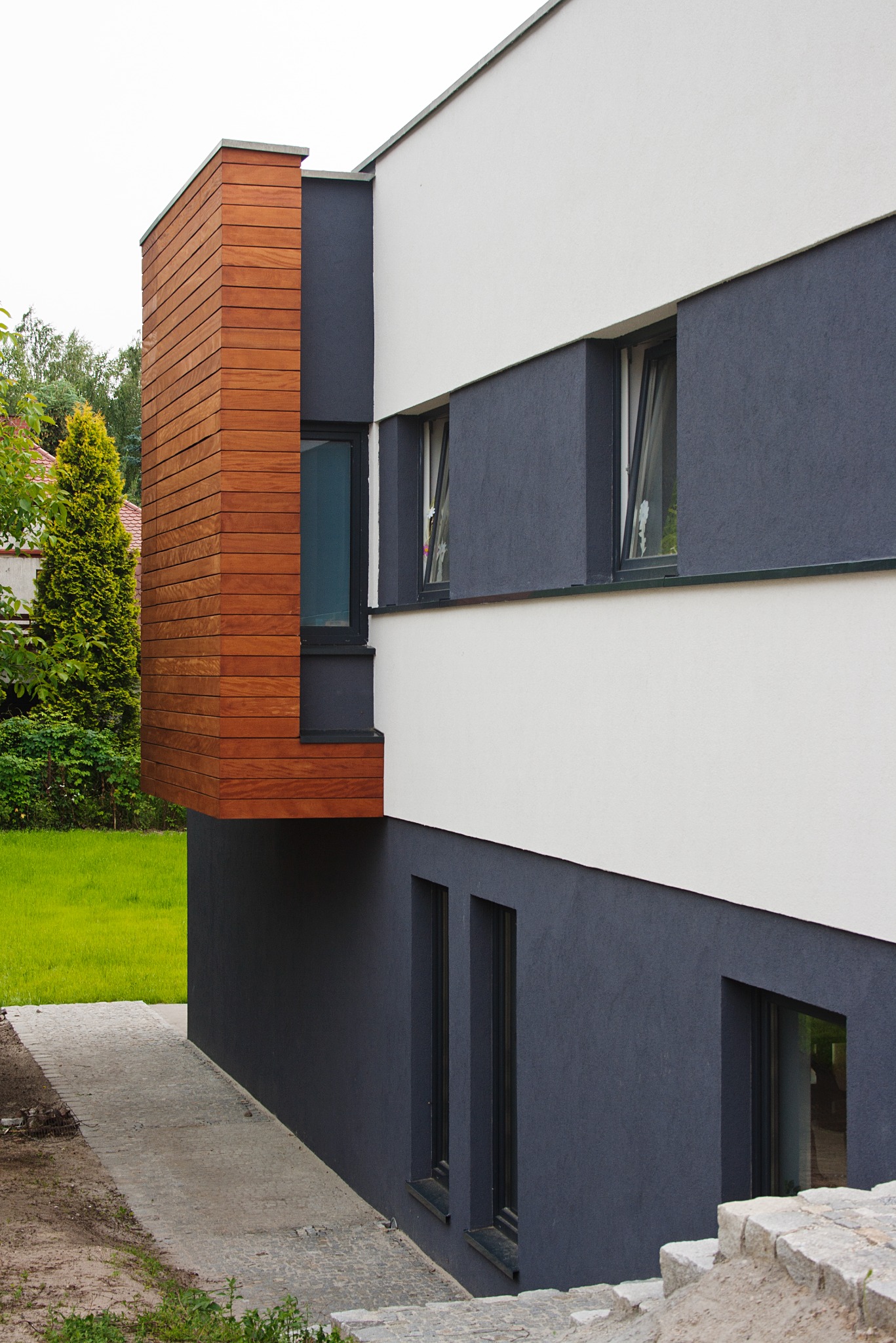 Fragment nowoczesnego budynku jednorodzinnego z drewnianą elewacją na wystającym elemencie, szara dolna część i biała górna część, widoczne okna i schody z kostki brukowej