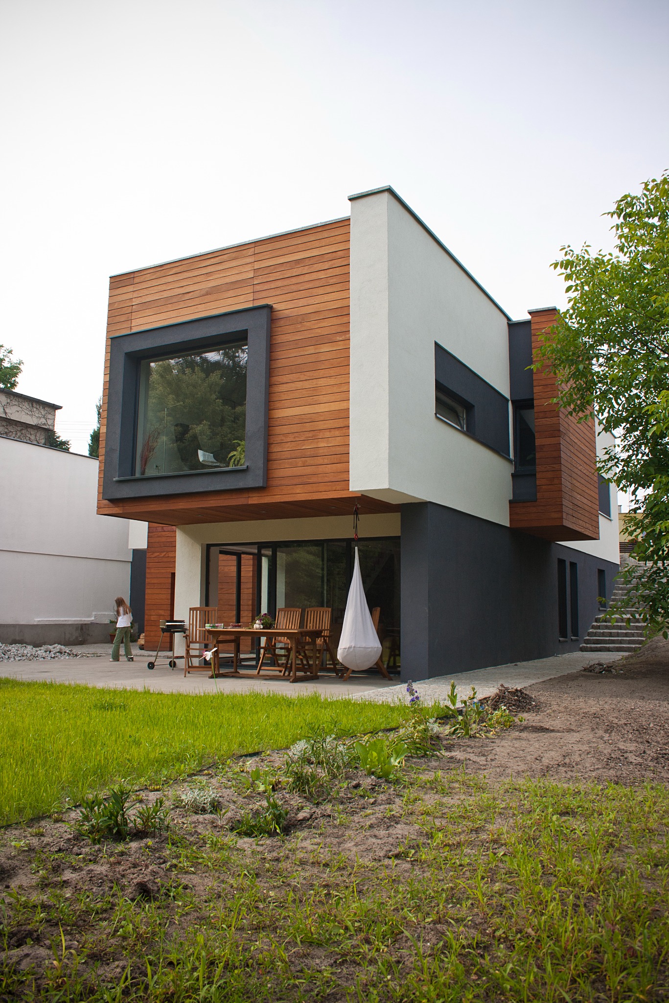 Nowoczesny budynek jednorodzinny w Poznaniu z drewnianą elewacją, dużymi oknami i tarasem z meblami ogrodowymi oraz wiszącym fotelem kokon. W tle widoczna kobieta przy grillu.