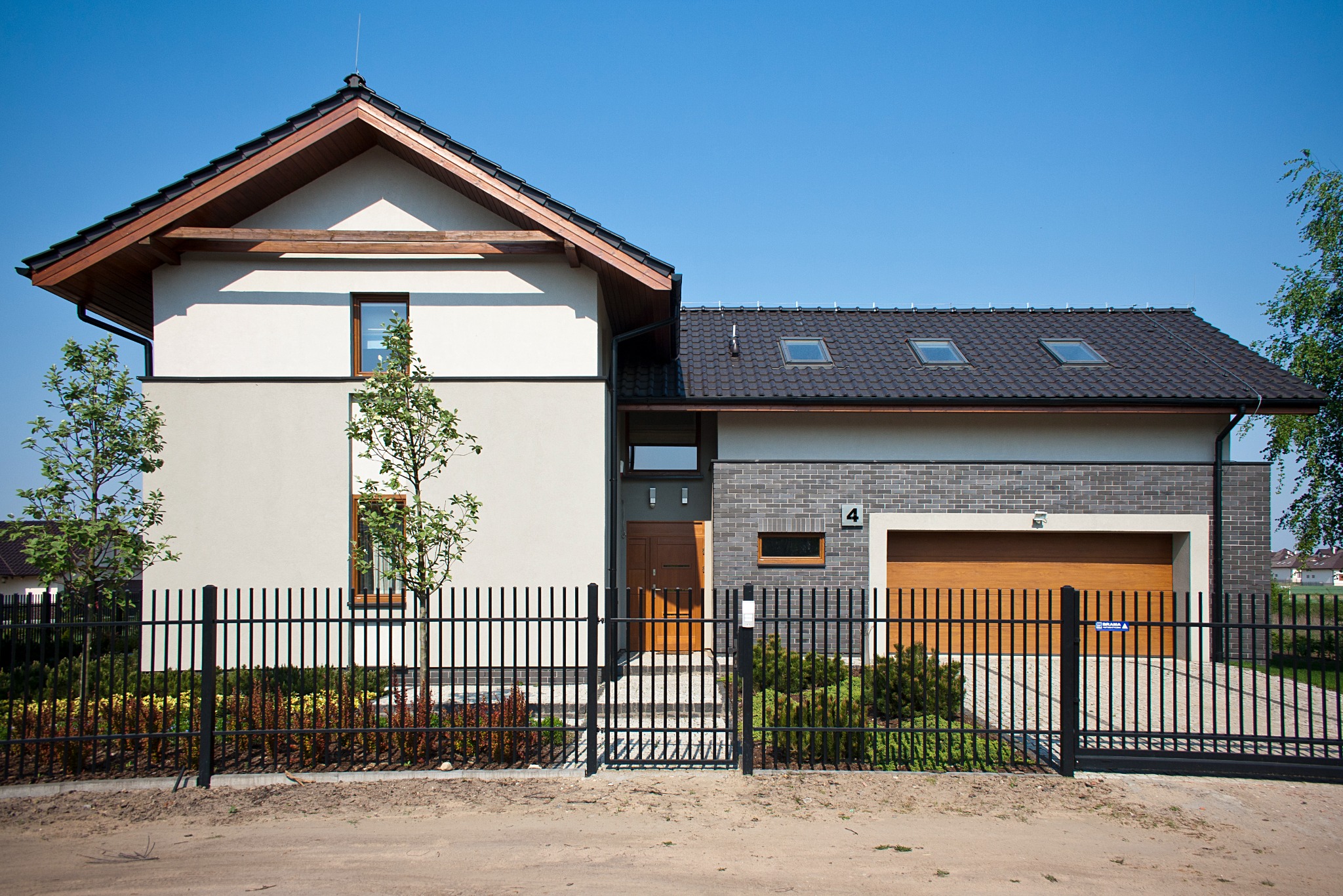 Nowoczesny dom jednorodzinny z dwuspadowym dachem i garażem, elewacja w odcieniach bieli i szarości, czarne metalowe ogrodzenie z furtką, niewielki ogródek przed domem, błękitne niebo w tle.