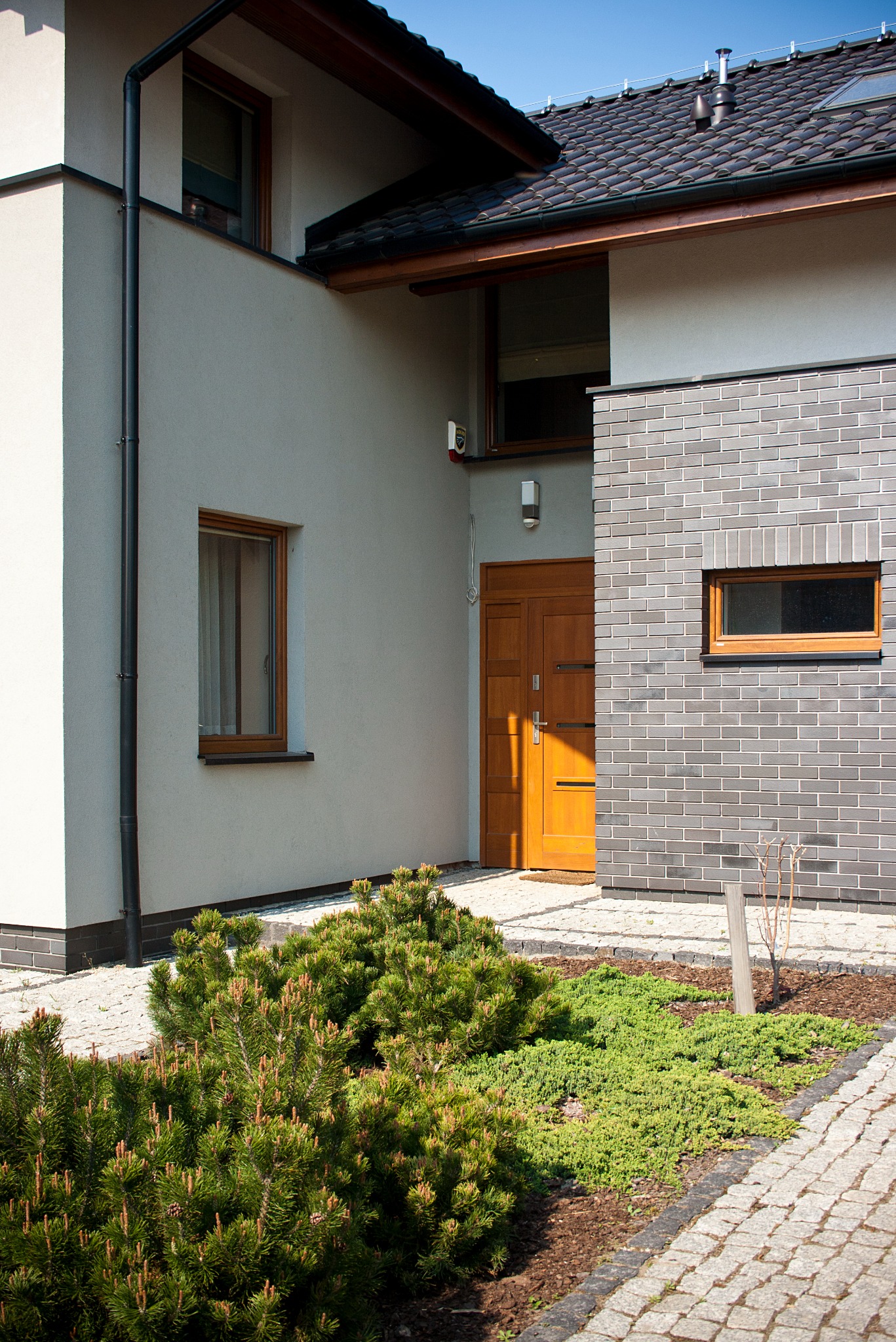 Nowoczesny dom jednorodzinny z elewacją z szarej cegły, drewnianymi oknami i drzwiami wejściowymi, otoczony zielenią i brukowanym chodnikiem.