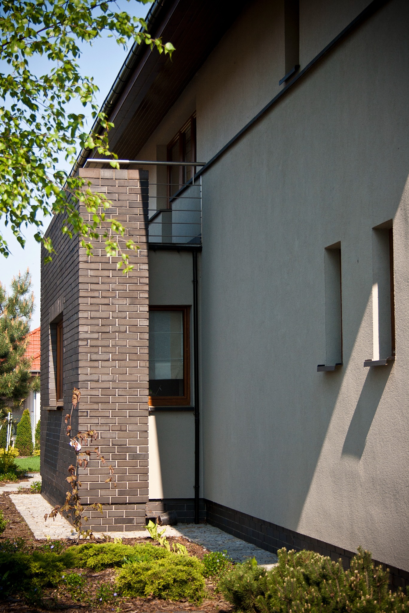 Fragment elewacji domu z jasnym tynkiem i ciemną cegłą klinkierową, mały balkon z metalową barierką, widoczne małe okna i fragment ogrodu z roślinnością.
