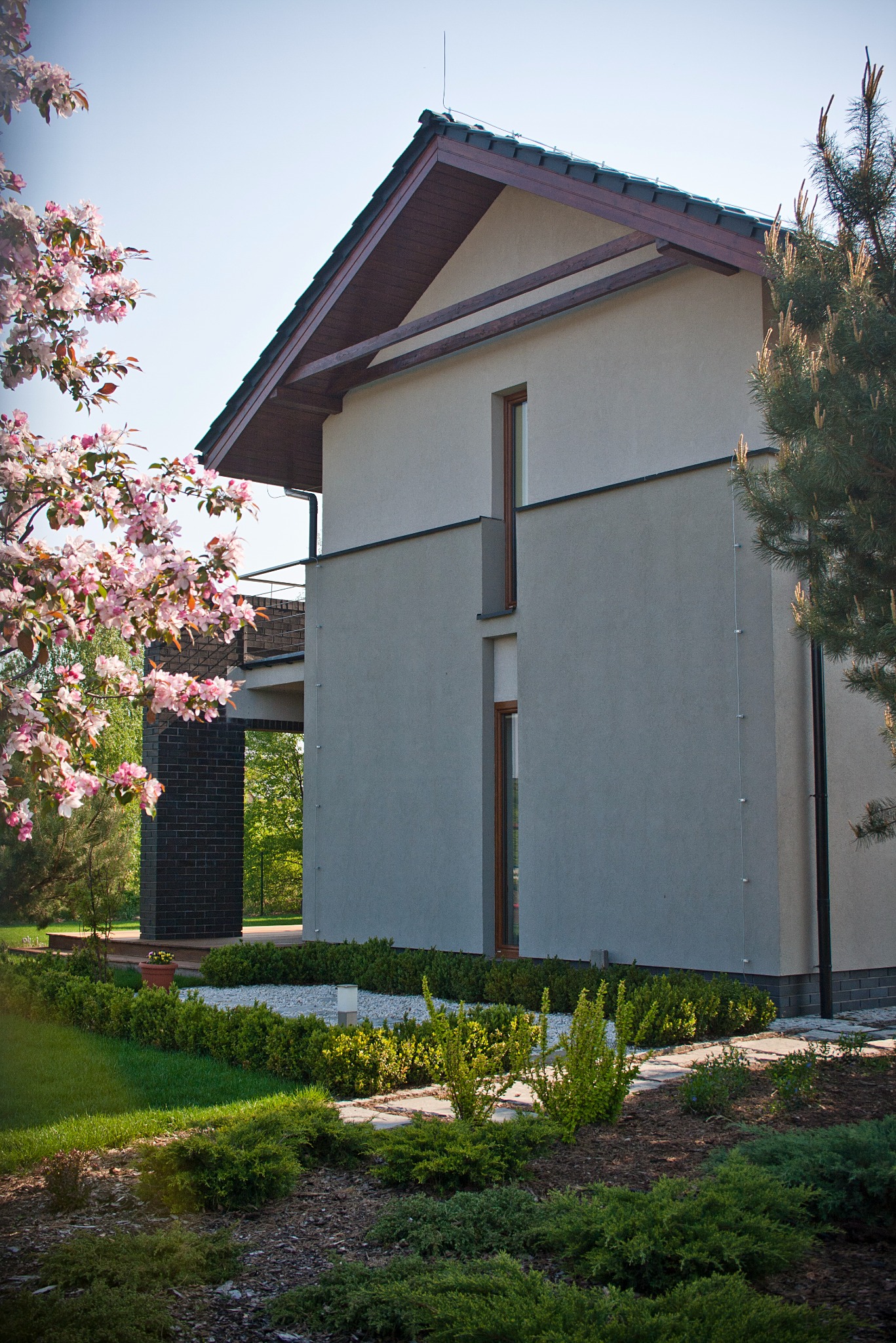 Nowoczesny dom z szarą elewacją, widok z ogrodu z kwitnącym drzewem i zadbanym trawnikiem, minimalistyczna architektura z elementami drewna.
