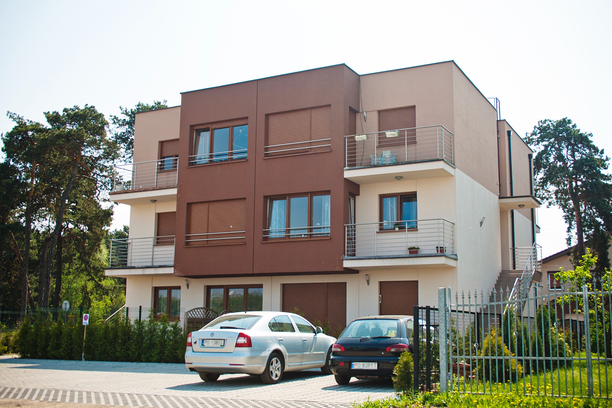 Trzykondygnacyjny budynek mieszkalny z balkonami, brązową i beżową elewacją, zaparkowane dwa samochody: srebrny i czarny.