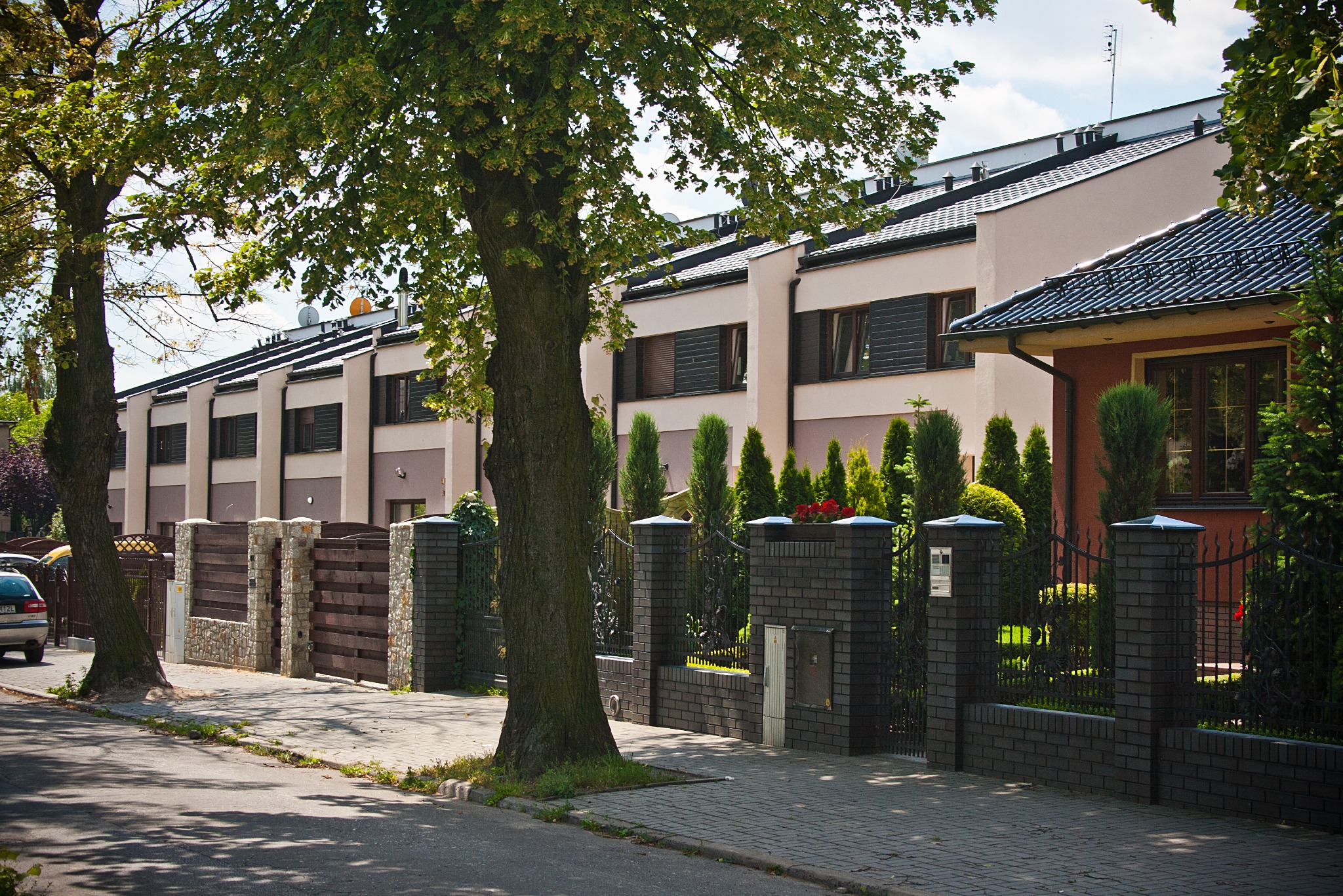 Szereg domów jednorodzinnych z ciemną stolarką okienną i ogrodzeniem z cegły klinkierowej, widok z ulicy w słoneczny dzień.