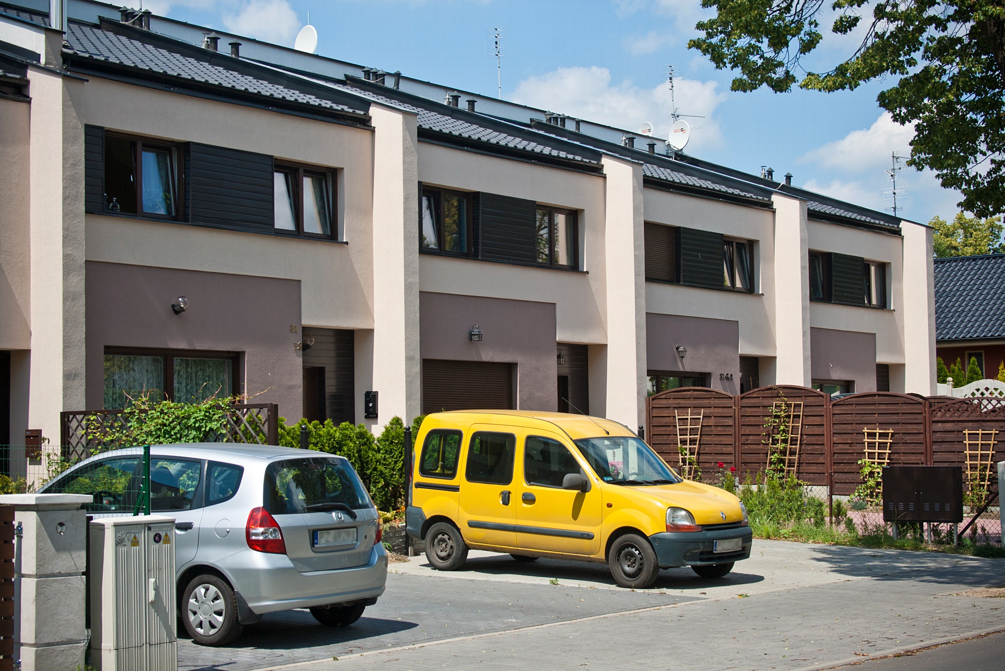 Szereg identycznych domów jednorodzinnych z płaskimi dachami, z zaparkowanymi samochodami: srebrnym hatchbackiem i żółtym vanem.