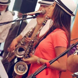 Kobieta w koralowej sukience gra na saksofonie podczas występu na żywo, w tle pozostali muzycy z saksofonami i mikrofonami, wszyscy w kapeluszach.